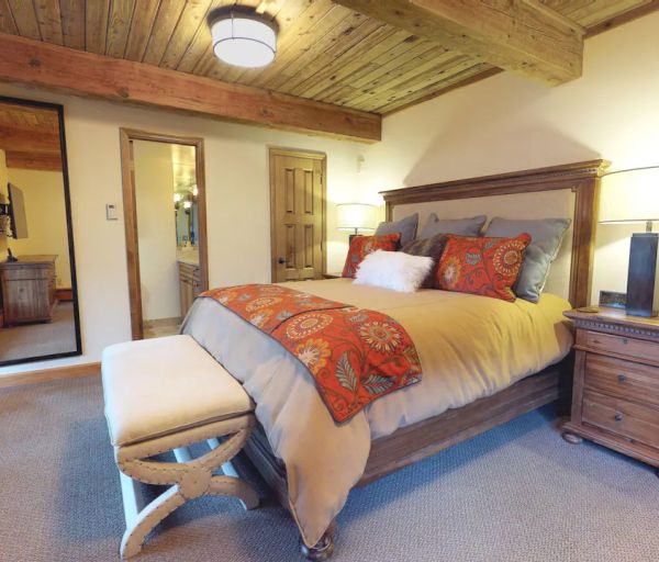 A cozy bedroom with a wooden ceiling, large bed, cushioned bench, bedside tables, lamps, a mirror, and a doorway leading to another room.