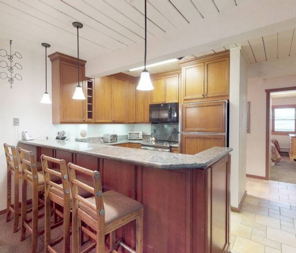 A kitchen with wooden cabinets, bar stools at a counter, pendant lights, and an adjacent hallway leading to other rooms.