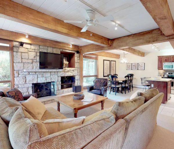 A cozy living room with wooden beams, a sectional sofa, a stone fireplace, TV mounted above it, dining area, and a kitchen with stainless steel appliances.
