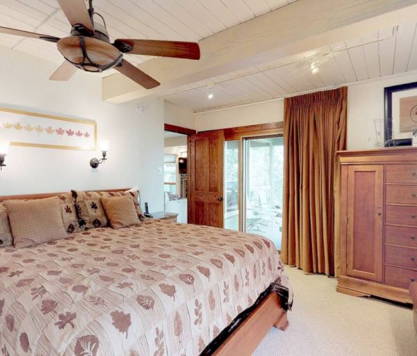A cozy bedroom with a large bed, ceiling fan, wooden furniture, art on wall, and sliding door leading outside.