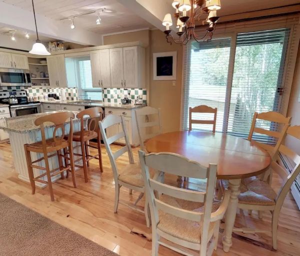 The image shows a cozy kitchen and dining area with wooden flooring, a round dining table with five chairs, a kitchen island, and modern appliances.
