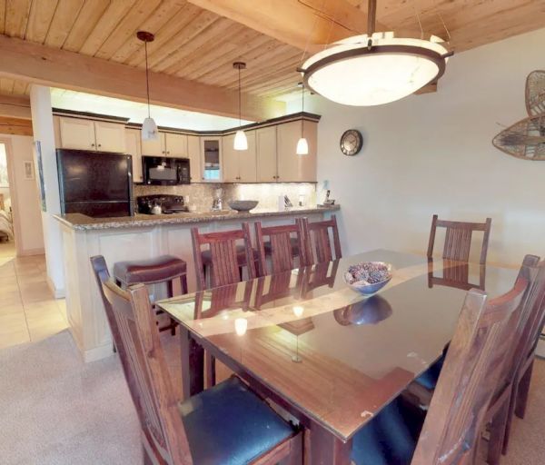 The image shows a cozy dining area with a wooden table and chairs, adjacent to a kitchen. A snowshoe decorates the wall to the right.