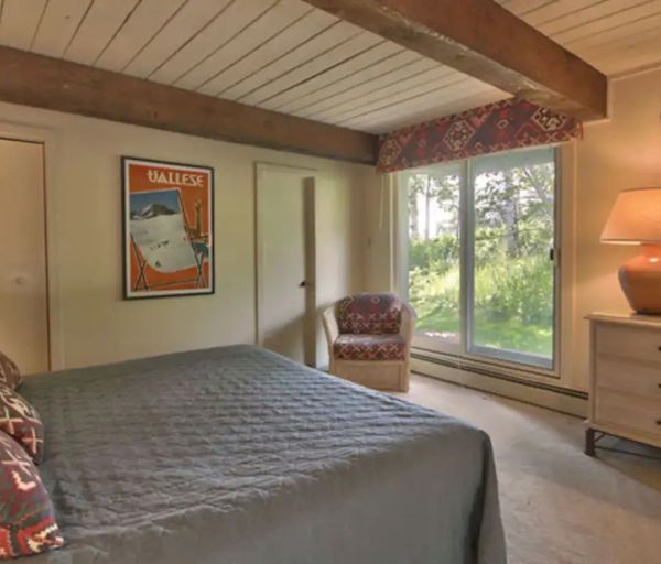 The image shows a cozy bedroom with a double bed, patterned pillows, wooden beams on the ceiling, a dresser with a mirror, and a large window.