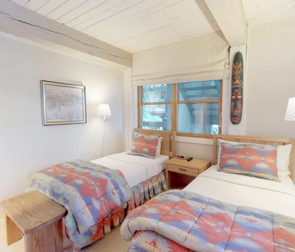 The image shows a cozy bedroom with two single beds, wooden furniture, and colorful bedding. A window allows natural light to enter.