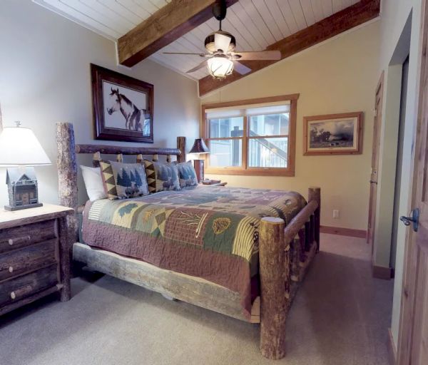 A cozy bedroom features a rustic wooden bed, matching dresser, bedside lamp, and framed paintings. Beige walls and a ceiling fan complete the setting.