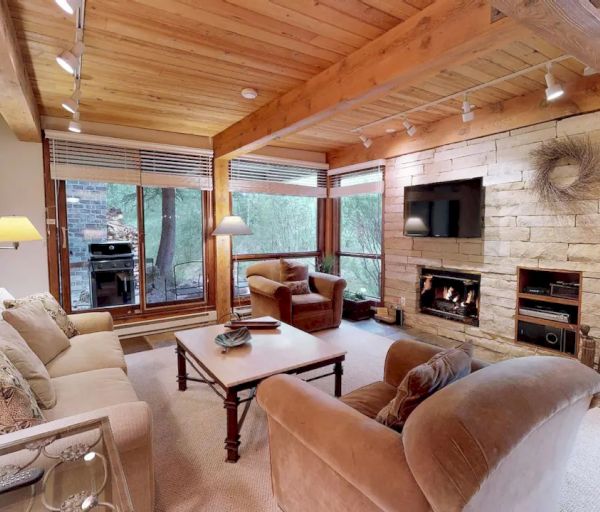 A cozy living room with wooden beams, a stone fireplace, a wall-mounted TV, large windows, and various seating options around a coffee table.
