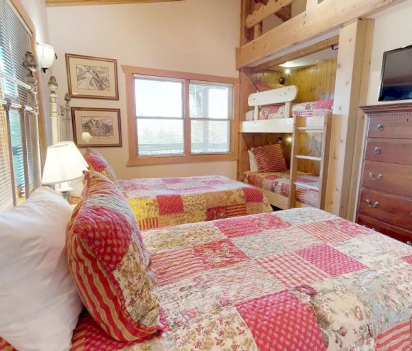 A cozy bedroom features two twin beds with patchwork quilts, a set of bunk beds, a wooden dresser with a TV on top, and framed pictures on the walls.