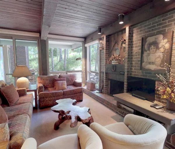 This image depicts a cozy living room with wooden ceilings, a sofa set, a rustic coffee table, and a fireplace, offering a view of the outdoors.