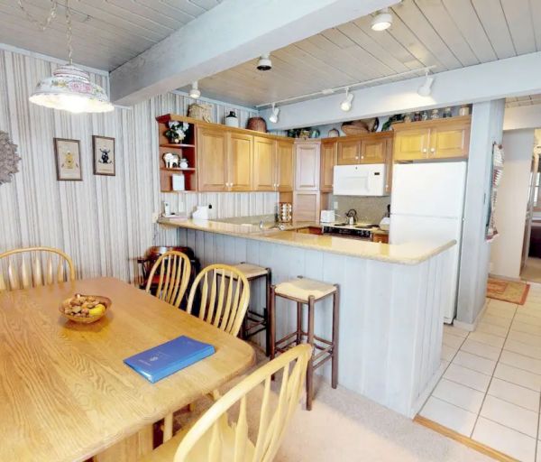 This image shows a cozy dining area and kitchen. The dining table has six chairs, and the kitchen features wooden cabinetry and a white fridge.