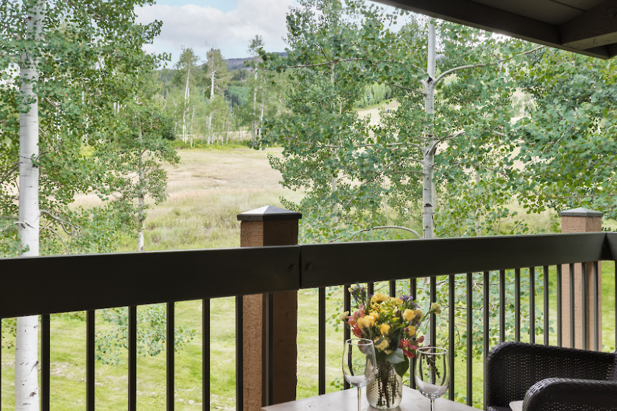 The image shows a balcony view with a flower vase, overlooking green trees and a grassy landscape in a serene setting.