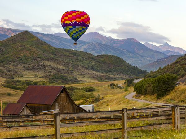 A colorful hot air balloon floats over a rural landscape with a barn, hills, and a winding road in the background.