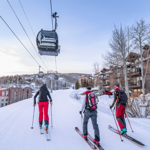 Three people are skiing uphill towards a ski lift in a snowy landscape, with buildings and trees in the background at dawn.