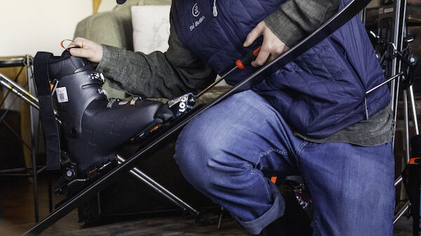 A person kneels while adjusting a ski boot, surrounded by ski equipment in a cozy indoor setting, with snowshoes hanging on the wall behind them.