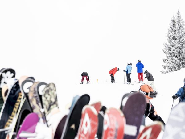 A group of skiers on a snowy slope with trees in the background, and a row of skis standing upright in the foreground.