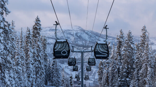 Snowy landscape with gondola ski lifts, pine trees, and mountains in the background. The lifts are suspended and heading up the slope.