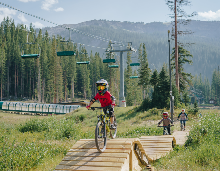 Three people are riding bikes on a wooden ramp in a scenic area with trees and mountains in the background, and ski lifts overhead, ending the sentence.