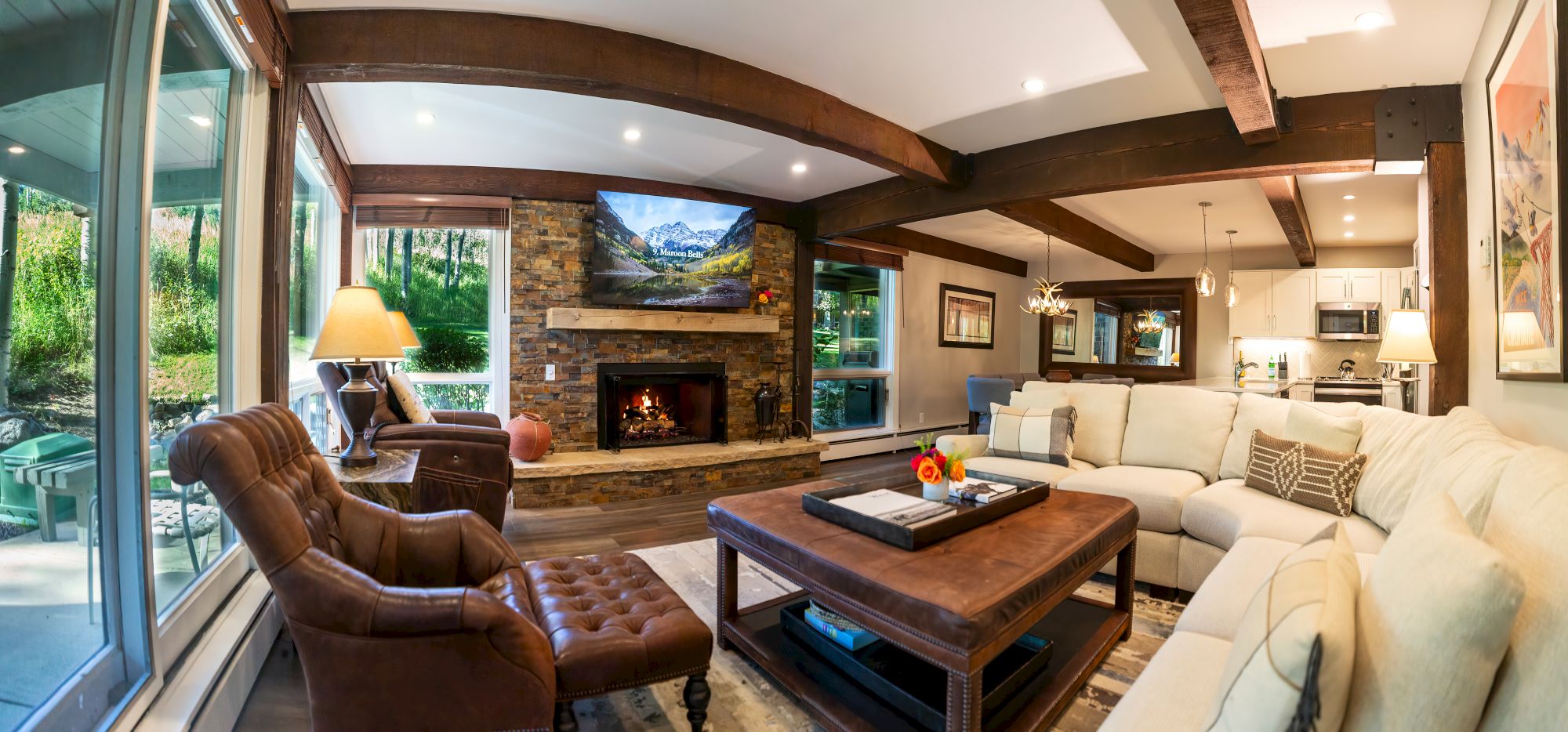 A cozy living room with a large window, fireplace, brown leather chairs, a beige sectional sofa, flat-screen TV, and a rustic wooden coffee table.