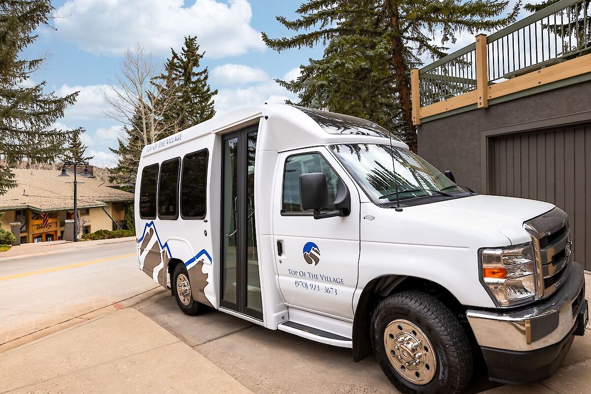 A white passenger van parked on a driveway near a building, with open side door and blue logo on the side, trees in the background, sunny day.